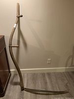 Full view of the antique wooden scythe showing the length of the wooden handle with two grips, standing against the wall.