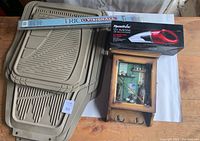Photo showing the beige automotive floor mats stacked, boxed RoadVac 12V auto vacuum, Trico Visionelle windshield wiper blade in packaging, and rustic wooden key holder with clock and hooks on a wooden table.