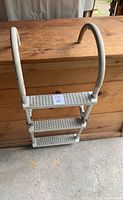 Front view of white metal boat ladder with three perforated steps and curved handrails against wooden background.