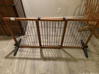 Full view of the dog gate extended, showing wood frame, metal wire grid, plastic or metal feet, and latch mechanisms (two broken). Scratches visible on wood frame.