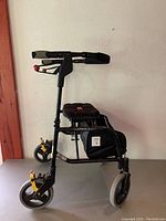 Side view of black folding walker with padded seat and storage bag, showing the three wheels, hand brakes, and frame structure.