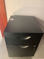 Front and side view of small black metal file cabinet with two drawers and lock on bottom drawer, placed on metal surface.