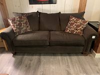 Front view of the brown upholstered sofa with three seat cushions and two multicolor patterned throw pillows.