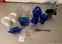 Full view of all glass items including white tumblers, blue glass cups, bowls, and small square glass dish grouped on table.
