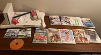 Photo showing Nintendo Wii console, sensor bar, two Wii Remote controllers (one with pink cover), balance board, and twelve Wii game cases on a wooden surface.