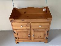 Full frontal view of the wooden dry sink showing two drawers with white ceramic knobs and double door cabinet with black hinges and white knobs. Top edge has scalloped decorative panel.