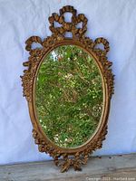 Full front view of ornate Renaissance style gold-colored plastic framed wall mirror with oval glass reflector, showing detailed floral and ribbon decoration on frame.