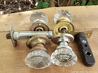 Photo of two vintage 1930s glass doorknobs with attached metal locksets showing the front side with clear faceted glass knobs and brass elements on a wooden surface outdoors.
