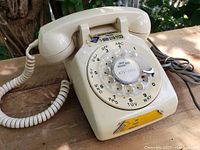 Ivory rotary dial phone showing left side and coiled handset cord on a wooden surface outdoors with green foliage in the background.