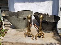 Pair of vintage galvanized coal scuttles with handles and brass andirons placed on a wooden surface.