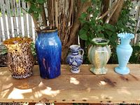 Five vintage vases displayed on wooden surface outdoors under tree shade. Vases vary in size, style and color.