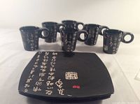 Photo of six black ceramic mugs featuring white Asian script and red stamps, arranged behind one square plate with matching calligraphy