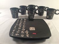 Front view of six black mugs and black square plates with white Asian calligraphy and red seal designs.
