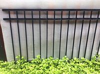 Black metal fence panel with vertical bars and horizontal rails resting against a wall and surrounded by greenery.