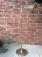 Full view of brass piano floor lamp against brick wall, showing round base, vertical pole, and rectangular shade.
