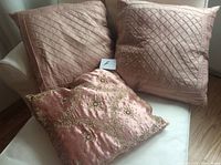 Photo showing two large pink cushions with diagonal stitched pattern and a smaller pink cushion with gold beaded embroidery placed on a white chair.