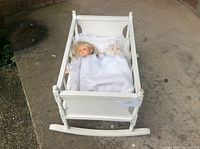 White wooden doll cradle with bedding and doll lying inside, shown from front angle on concrete floor