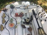 Top down view of all thirteen garden ornaments on metal sticks arranged on a white table outdoors, showing a variety of decorative designs including butterfly, frog, bird, spider, leaves, fleur-de-lis, and lantern.