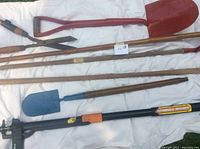 Photo showing group of garden tools on white cloth including two large red shovels, a blue shovel, an extendable black metal garden tool with wooden handle, and wooden poles.