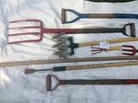 Various garden tools (pitchforks, cultivators, shovels) laid on a white sheet for sale.
