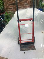Small red moving cart with a rectangular base and two large wheels, shown upright against a plastic-covered background.