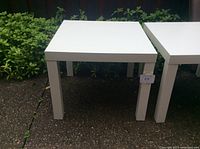 First image showing one of the two white square plastic tables outdoors on a concrete surface with plants in the background.
