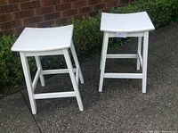 Two white painted wooden bar stools shown from above and side angles outdoors on pavement near foliage and a brick wall.