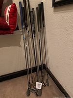 Photo showing full set of nine golf clubs standing against wall, various grips and heads visible
