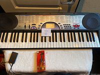 Casio electronic keyboard with built-in speakers and power adapter plug visible on the left side, placed on white surface.