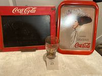 Photo of Coca Cola chalkboard, vintage tray with woman in hat, and Coca Cola fountain glass.