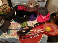 Photo shows assortment of hats including baseball caps and bucket hats, Bell helmet, swim goggles, and pool inflatables with smash ball set in foreground