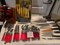 Overall view of red handled silverware, wooden knife blocks, assorted cutlery knives with black handles and orange protective cover, and Cambridge flatware box in the background