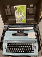 Blue Smith Corona electric typewriter with Sterling Cartridges badge, black keys, and light blue casing, placed inside its brown hard plastic case. Manual included.