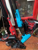 Photo showing red Dirt Devil stick vacuum, blue Bissell stick vacuum, black portable car vacuum, and small red shampooer.