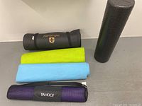 Four rolled yoga mats and one black foam roller arranged on floor against wall. Mats are black, purple, blue, and green. Purple mat is inside a mesh carrying case.