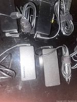 Photo of four black Lenovo branded AC power adapters with attached cables and slim tip connectors, plus power cords coiled beside them, all new condition.