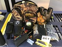 Full hunting equipment lot arranged on a black metal surface showing camouflage bag, headlamp, rope, sheaths, and multi-tool.