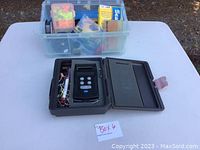 Photo showing the digital fishing weigh scale in its open protective case and a plastic container filled with assorted fishing tackle items on a white table.