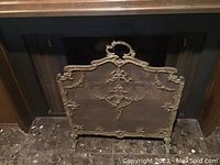 Front view of the fireplace screen showing brass ornamentation over wire mesh with decorative ribbons, rosettes, and foliage.