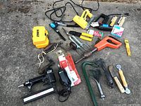 Overview of various hand and power tools laid out on the ground including tape measures, stapler, electric tools, screwdrivers, wrenches, and garden tools.