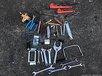 Photo showing overall assortment of hand tools laid out on dark surface, highlighting pipe wrenches, pliers, wrenches, and scraper.