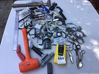 Photo showing a wide assortment of small hardware including clamps, rubber mallet, hammer, scissors, and assorted metal parts spread out on a white table with a ruler alongside.