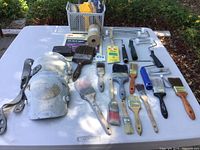 Layout of knee pads, paint brushes, roller handles, and painting trays on a table outdoors.