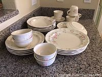 Photo showing stacked dinnerware including dinner plates, mugs, bowls, and smaller plates, with floral Tea Rose pattern visible on items.
