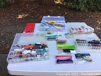 Photo showing multiple small plastic tackle boxes with assorted soft plastic fishing lures and packaged lures spread on a white table outside.