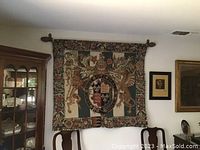 Wall-mounted loom woven tapestry depicting the Royal Arms of England with heraldic shield, griffins, and floral border. Shown hanging on a wooden rod with finials in a home setting beside wooden cabinet and framed pictures.