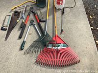Yard tools including rakes, sprayers, clippers, and saws spread out on concrete ground