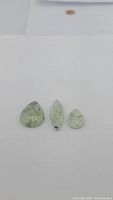 Three rutilated prehnite gemstones placed side by side: one pear-shaped and two marquise cabochon cuts, showing their translucent light green color with needle-like rutile inclusions.