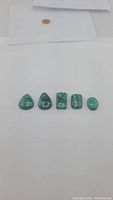 Five green malachite cabochons laid in a row showing various shapes and natural patterns against a white background