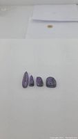 Four polished natural purple charoite gemstones placed in a row on white background, showing size and shape differences.